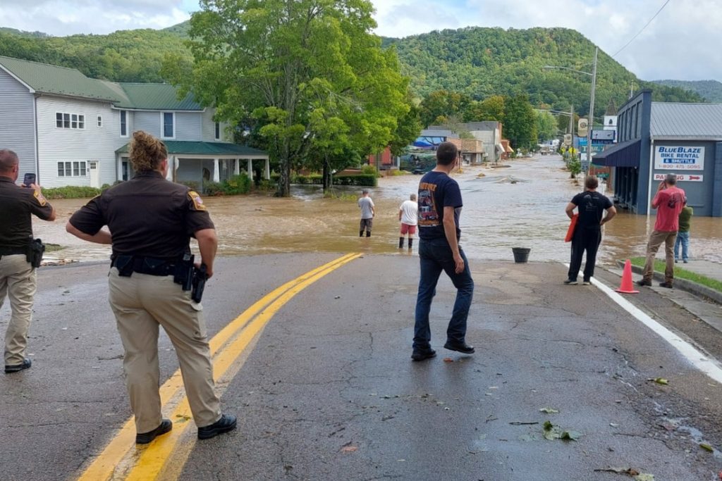 Law enforcement and residents stand at the edge of a flooded main street in Damascus, Virginia, as muddy water flows past downtown buildings.