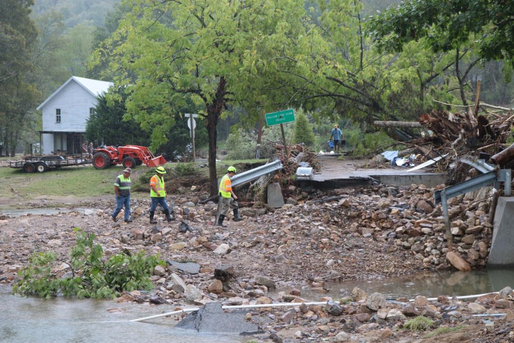 Road crews in safety vests inspect the washed-out Route 725 bridge in Taylors Valley, amid debris, damaged guardrail, and floodwater near a farmhouse.